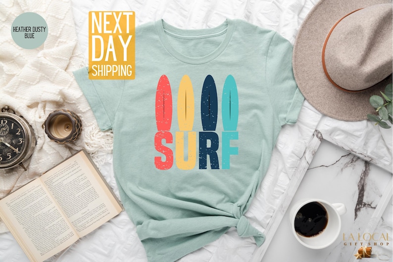 May include: A light blue t-shirt with a retro-style graphic of four colorful surfboards and the word "SURF" in bold letters.