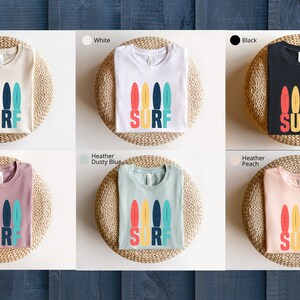May include: A set of ten t-shirts in various colors featuring a graphic of colorful surfboards with the word "SURF" printed below. The t-shirts are displayed on a wooden background.