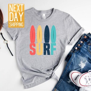 May include: A heather light grey t-shirt with a retro-style graphic of four colorful surfboards and the word "SURF" in large, bold letters.