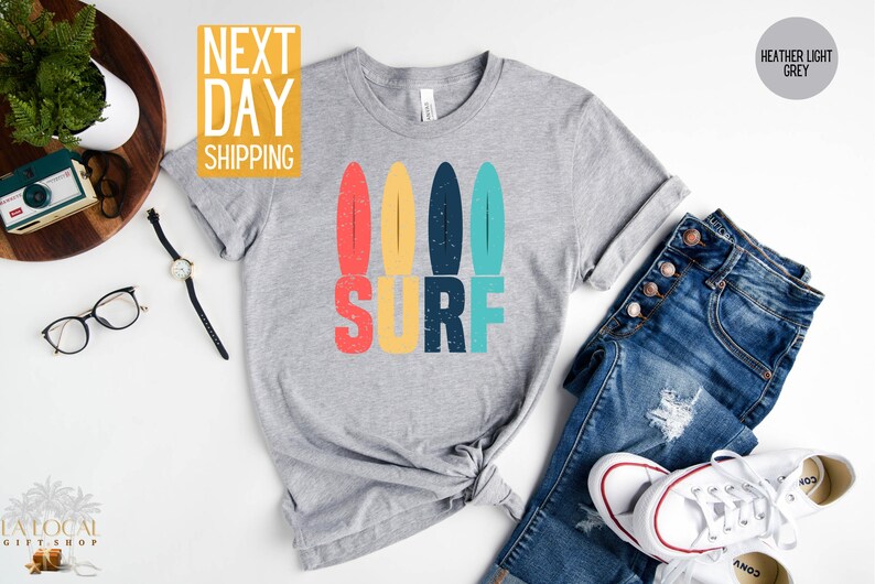 May include: A heather light grey t-shirt with a retro-style graphic of four colorful surfboards and the word "SURF" in large, bold letters.
