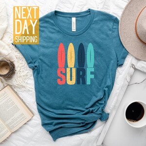 May include: A teal blue t-shirt with a graphic of four colorful surfboards and the word "SURF" in colorful retro lettering.