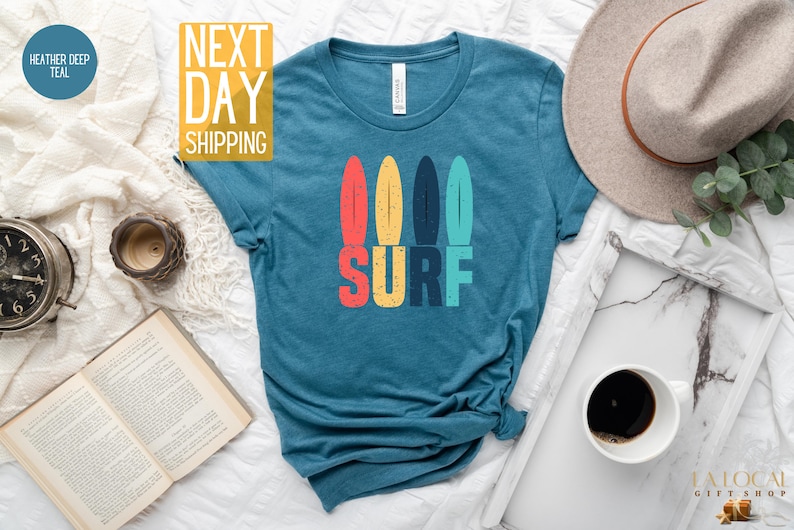 May include: A teal blue t-shirt with a graphic of four colorful surfboards and the word "SURF" in colorful retro lettering.