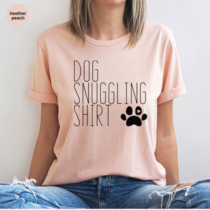 May include: A light pink t-shirt with the text "DOG SNUGGLING SHIRT" and a black paw print with a small white heart in the center.