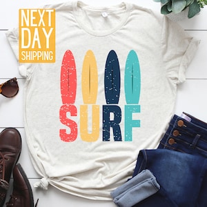 May include: A white t-shirt with a vintage-style graphic of four colorful surfboards and the word "SURF" in bold letters. The graphic has a distressed look, giving it a retro feel.