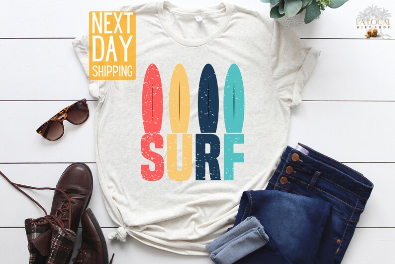 May include: A white t-shirt with a vintage-style graphic of four colorful surfboards and the word "SURF" in bold letters. The graphic has a distressed look, giving it a retro feel.