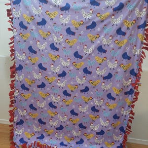 May include: A lavender fleece blanket featuring a repeating pattern of colorful chickens in white, yellow, blue, and navy. The blanket has a red fringe border, adding a decorative touch to this cozy home textile.