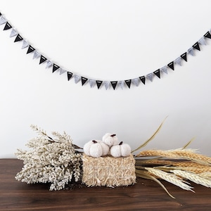 May include: A festive autumn display featuring a garland of black and silver triangle flags. Three small, white knitted pumpkins rest on a square bale of hay. The arrangement includes dried wheat and a bouquet of white flowers, all set on a dark wooden surface.