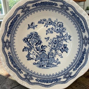 May include: A white ceramic bowl featuring a blue transferware pattern. The design includes a central floral motif with blooming flowers and a small butterfly, surrounded by a decorative geometric and floral border along the scalloped rim.