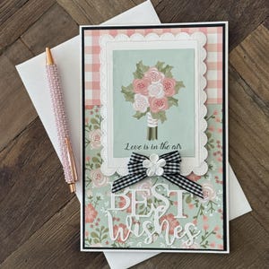 May include: Greeting card with a floral bouquet illustration, the text "Love is in the air", and the words "Best Wishes". The card has a gingham and floral pattern, a black and white bow, and a pink pen.