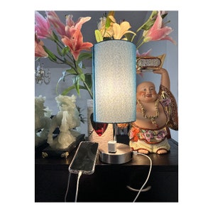 May include: A blue lampshade with a white fabric shade sits on a table with a figurine of a smiling Buddha. The lamp is plugged in and has a white cord.