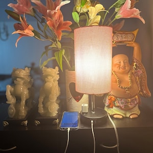 Puede incluir: Una lámpara blanca con una pantalla de tela rosa se encuentra sobre una superficie de madera oscura. Dos estatuas de jade verde están a cada lado de la lámpara. Un teléfono móvil está conectado al puerto USB de la lámpara. Una pequeña figura de porcelana de un Buda sonriente está a la derecha de la lámpara.