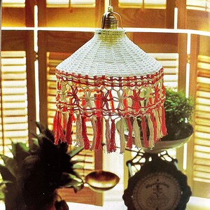 May include: A white crocheted lampshade with red and white fringe. The lampshade is hanging from a ceiling fixture and is surrounded by plants.