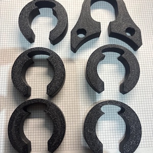 May include: A collection of six black, textured, 3D-printed parts. The parts include four C-shaped pieces and two pieces with a curved top and two holes. The items are arranged on a grid-lined surface.