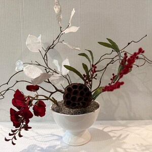 May include: A white ceramic pedestal bowl containing a floral arrangement with dark red flowers, white translucent leaves, a brown dried lotus seed pod, green leaves, and red fuzzy berries, all set against a plain white background.