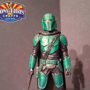 K&ouml;nnte beinhalten: Eine Mandalorianer-Actionfigur in Gr&uuml;n und Schwarz, die auf einer kreisf&ouml;rmigen Basis mit einem gr&uuml;nen Design steht. Die Figur hat einen Helm, eine R&uuml;stung und eine Waffe. Das Logo "TONY & CHRIS CRAFTS ESTD. 2020" befindet sich oben links.