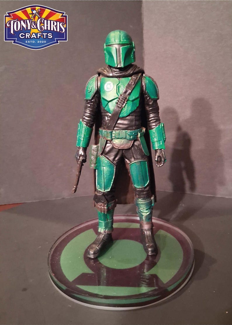K&ouml;nnte beinhalten: Eine Mandalorianer-Actionfigur in Gr&uuml;n und Schwarz, die auf einer kreisf&ouml;rmigen Basis mit einem gr&uuml;nen Design steht. Die Figur hat einen Helm, eine R&uuml;stung und eine Waffe. Das Logo "TONY & CHRIS CRAFTS ESTD. 2020" befindet sich oben links.
