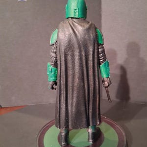 K&ouml;nnte beinhalten: Eine gr&uuml;ne und schwarze Actionfigur einer Figur mit Helm und Umhang, die auf einer runden Basis mit gr&uuml;nem und schwarzem Design steht. Die Figur h&auml;lt ein kleines, braunes Objekt. Das Logo "TONY & CHRIS CRAFTS ESTD. 2020" befindet sich in der oberen linken Ecke.