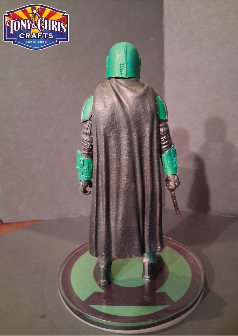 K&ouml;nnte beinhalten: Eine gr&uuml;ne und schwarze Actionfigur einer Figur mit Helm und Umhang, die auf einer runden Basis mit gr&uuml;nem und schwarzem Design steht. Die Figur h&auml;lt ein kleines, braunes Objekt. Das Logo "TONY & CHRIS CRAFTS ESTD. 2020" befindet sich in der oberen linken Ecke.