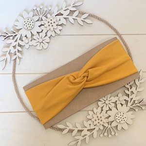 May include: A mustard yellow knotted headband made of soft fabric. The headband is displayed on a brown background with white wooden flowers.