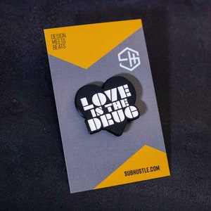 Op de afbeelding: Zwarte en witte emaille pin in de vorm van een hart met de tekst "LOVE IS THE DRUG" in een retro-lettertype. De pin is op een grijze en gele kaart met de tekst "DESIGN MEETS BEATS" en "SUBHUSTLE.COM".