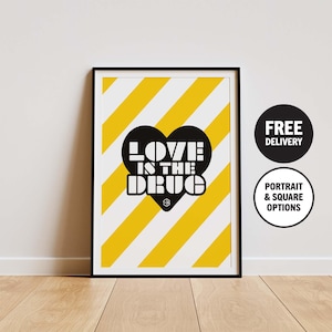 Puede incluir: Impresión artística enmarcada con borde negro. La obra presenta una forma de corazón negro con las palabras "LOVE IS THE DRUG" en blanco, sobre un fondo a rayas amarillas y blancas. La impresión se muestra en una pared blanca.