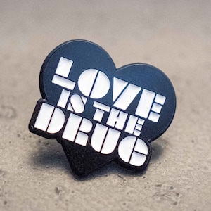 Puede incluir: Pin negro en forma de corazón con texto blanco que dice "LOVE IS THE DRUG".
