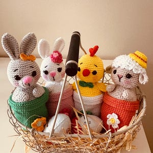 Peut inclure: Un panier en fil de fer rempli de jouets de Pâques crochetés. Le panier contient un lapin gris, un lapin blanc, un poussin jaune et un mouton. Chaque jouet porte un petit accessoire. Le panier contient également des œufs décoratifs et de la paille.