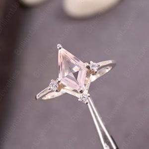 May include: A silver ring with a light pink, kite-shaped gemstone at its centre. The ring is embellished with small, round, clear gemstones. The ring is held by tweezers. The ring is a piece of jewellery.