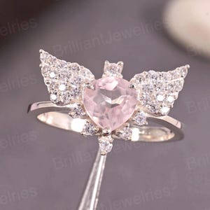 May include: A silver ring featuring a heart-shaped, light pink gemstone. The ring is adorned with small, clear gemstones and wing-like embellishments on either side of the heart. The ring is held by a pair of tweezers.