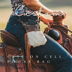 May include: A brown leather clip-on cell phone bag with a fringe and a white fur flap. The bag is attached to a belt and is being worn by a person riding a horse. The text "Custom Clip On Cell Phone Bag" is visible at the bottom of the image.