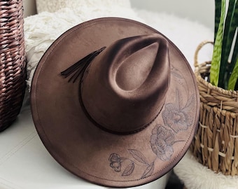 May include: A brown suede fedora hat with a floral design and a tassel. The hat has a wide brim and a tall crown.