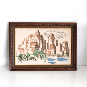 May include: Embroidered cityscape art in a dark wood frame. The artwork depicts a skyline with brown, black, and tan buildings, green trees, and a blue water feature. A light blue cloud is stitched in the sky.