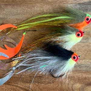 Peut inclure: Trois leurres de pêche à la mouche avec des plumes orange, vertes et noires. Les leurres ont des yeux orange vif et sont disposés en rangée sur une surface en bois.