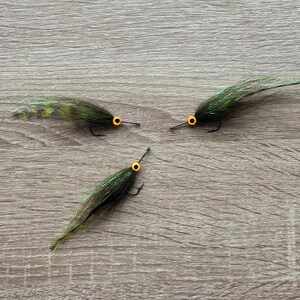 Peut inclure: Trois leurres de pêche à la mouche vert et noir avec des yeux jaunes. Les leurres sont liés avec des plumes et sont disposés en forme de triangle sur une surface en bois.