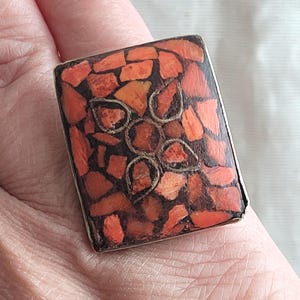 May include: A rectangular ring with a mosaic design. The ring's face is filled with small, irregularly shaped pieces in shades of orange and red, set against a dark background. A silver-coloured floral design is in the centre.