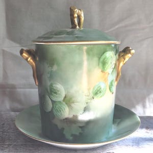 May include: A vintage porcelain jar with lid and matching saucer. The jar is painted with green and white gooseberries and leaves. It has gold-coloured handles and a gold accent on the lid. The jar sits on a matching saucer.