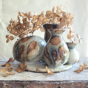 May include: A set of ceramic vases in different sizes and shapes, with a speckled brown and blue glaze. The vases contain dried, brown flowers, and are displayed on a light-coloured, rustic wooden surface.