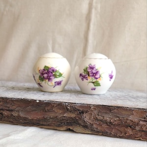 May include: Two white ceramic salt and pepper shakers with a floral violet design. The shakers have a rounded shape and small lids. The violet flowers are purple with green leaves. The shakers are on a wooden surface.