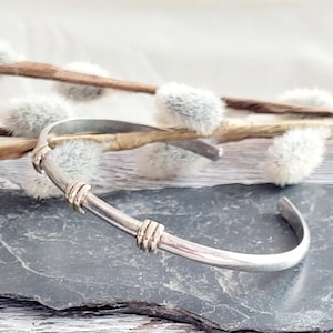 May include: A silver cuff bracelet with gold-coloured detailing. The bracelet has a minimalist design with a curved form. The gold accents are wrapped around the silver band. The bracelet is displayed on a dark grey surface with a blurred background.