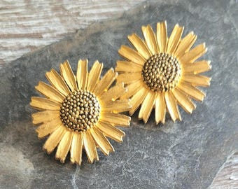 May include: A pair of gold-coloured daisy earrings. Each earring has a detailed flower design with a textured centre and radiating petals. The earrings are set against a dark, textured surface.
