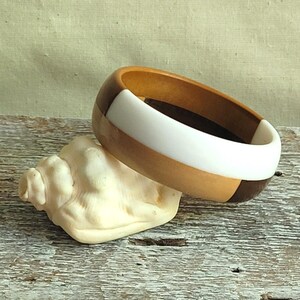 Peut inclure: Un bracelet jonc épais composé de segments alternés en bois clair, en bois foncé et en matériau blanc uni. Le bracelet présente une surface extérieure arrondie et polie ainsi qu'une conception en anneau large avec une finition lisse.