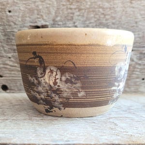 May include: A beige ceramic bowl with a dark brown band featuring a floral design. The bowl has a light brown rim and a textured, rustic appearance. The floral design is in a lighter brown colour, adding contrast to the darker band.