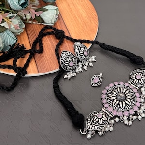 May include: A silver-toned necklace and earring set with a floral design and small white beads. The necklace has a black cord. The earrings and pendant have a teardrop shape. The set includes a pendant and earrings.