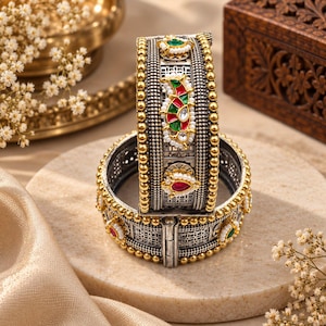 May include: A hinged metal bangle bracelet featuring an oxidized silver finish with gold-toned beaded edges. The surface is decorated with small white pearls and inlaid enamel accents in red and green, arranged in floral and paisley-inspired patterns.