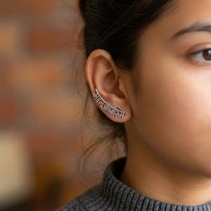 Puede incluir: Un pendiente de plata con el texto sánscrito "Aham Brahmasmi" curvándose a lo largo de la oreja. La joya se lleva en la oreja de una persona que lleva un jersey gris. El fondo es una pared de ladrillos borrosa.