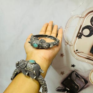 Può includere: Due braccialetti a forma di elefante color argento con dettagli turchesi. I braccialetti presentano dettagliati disegni di elefanti e sono esposti su una mano. I gioielli sono ambientati su uno sfondo neutro.