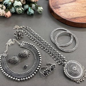 Puede incluir: Un surtido de joyas plateadas, que incluyen un collar, aretes, anillos, brazaletes y una pieza para la cabeza. Las joyas presentan intrincados diseños de cuentas y flores. Las piezas están dispuestas sobre una superficie gris, con un plato de madera y un arreglo floral en el fondo.