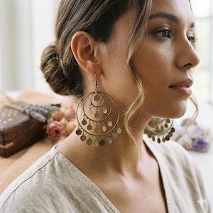 May include: Gold-toned hoop earrings with multiple concentric circles and dangling coin-shaped charms. The earrings are worn by a person with light skin and wavy hair. The background includes a wooden box and flowers.