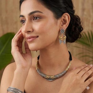 Puede incluir: Un conjunto de joyas plateadas y doradas con un collar, pendientes y pulsera. El collar tiene un dise&ntilde;o curvo con detalles intrincados y un cord&oacute;n negro. Los pendientes tienen una parte superior floral y un dise&ntilde;o en forma de coraz&oacute;n. La pulsera tiene un dise&ntilde;o a juego.