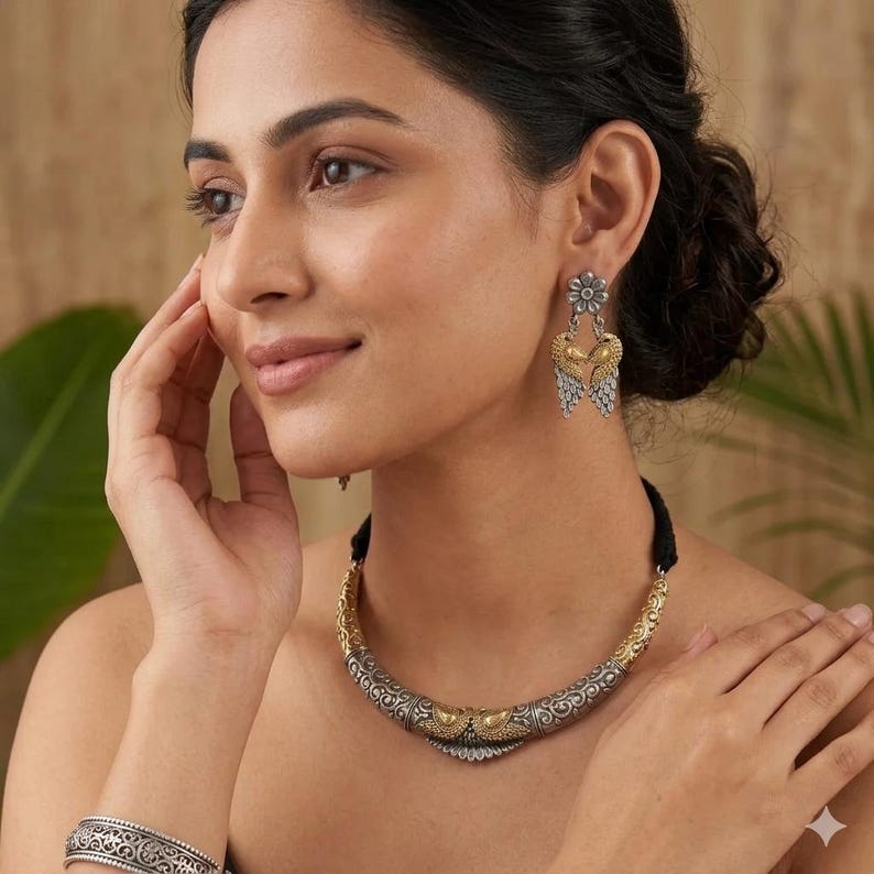 Puede incluir: Un conjunto de joyas plateadas y doradas con un collar, pendientes y pulsera. El collar tiene un dise&ntilde;o curvo con detalles intrincados y un cord&oacute;n negro. Los pendientes tienen una parte superior floral y un dise&ntilde;o en forma de coraz&oacute;n. La pulsera tiene un dise&ntilde;o a juego.